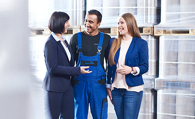Team members in a warehouse discussing, with two women in business attire and one in workwear.