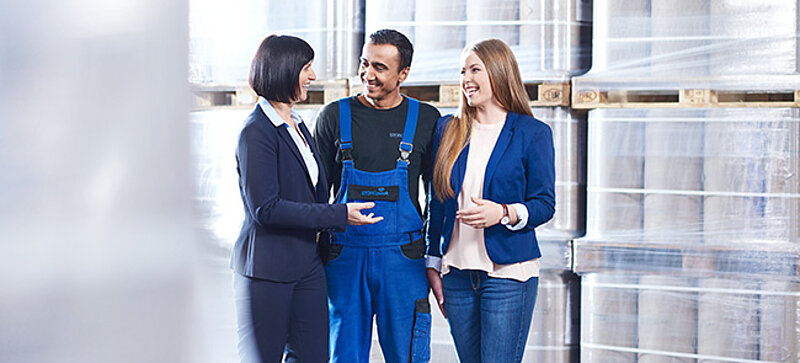 
		  Team members in a warehouse discussing, with two women in business attire and one in workwear.