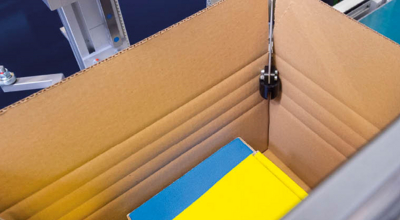 
				Cardboard box on a production line, where a machine is cutting the edges for precise packaging.