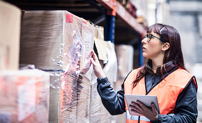 A woman waering a high-visibility vest standing in front of a warehouse rack containing boxes